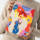 Person in beigem Strickpullover hält das bunt gestaltete Magazin „flow“ vor hellgrauem Hintergrund, Titelseite mit gelbem Schriftzug „flow“, Unterzeile „Das Magazin für persönliche Entwicklung, Kreativität & mehr Ruhe“ sowie zusätzlicher Aufschrift im blauen Kreis „DIY-Girlande & Sommerjournal“. Abgebildete Illustration zeigt eine Person in blauer Latzhose mit bunten Farbkleksen und Pinsel vor abstraktem, farbenfrohem Hintergrund.