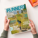 Titelblatt des Laufmagazins „Runner’s World“ mit einer laufenden, lächelnden männlichen Person in gelbem Shirt, blauer Shorts und Sonnenbrille auf einem sonnigen Bergpfad. Hauptüberschriften: „Das Geheimnis der Superschuhe“, „Warum bin ich nicht schneller?“, „Es gibt kein Alterslimit“, „So schläfst du besser – ein Leitfaden“, „Die Wunderläuferin“. Hände halten die Zeitschrift vor hellem Hintergrund, orangefarbenes Tablett im Eck.