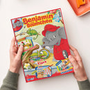 Kinderzeitschrift mit rotem Rand und dem Titel „Benjamin Blümchen“ in gelber Schrift. Auf der bunt illustrierten Vorderseite ist ein grauer Elefant mit roter Mütze und Hemd zu sehen, daneben ein Chamäleon in Grün- und Gelbtönen auf einem Ast und in einer Lupe. Textfelder: „Leckere Gemüsesnacks“, „So sehen Tiere“, „Doras Rechenpyramide“ und „Ein fliegendes Chamäleon“. Zwei Hände halten das Heft über einem hellen Tisch.
