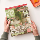 Zwei Hände in grünem Strickpullover halten eine Ausgabe der Zeitschrift „Wohnidee“ mit hellgrünem Titelbalken und dem Untertitel „Wir lieben unser Zuhause!“. Das Cover zeigt eine sommerliche Gartenszene mit gedecktem Tisch, rosa und grünen Kissen und Pflanzen im Hintergrund. Sichtbare Schlagzeilen u. a.: „Lieblingsplatz im Grünen“, „Gesund schlafen“, „Da blüht uns was!“ und „Eigene Ernte“.