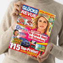Person in beigem Strickpullover hält das Magazin „Glücks Revue“ mit farbenfrohem Cover, gelben, roten und blauen Textfeldern. Titelthemen u. a.: „Ihr Glück liegt in Scherben“, „So senken Sie das Risiko für Diabetes“, „Lecker! Die besten Rezepte zum Schlemmen“, „115 Top-Preise“, dazu Fotos von einer blonden Frau, einem blauen Auto, Tortenstücken, einem tropischen Strand und einem Mann im Porträt.
