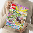 Person in beigem Strickpullover hält eine Ausgabe der Zeitschrift „Lisa Wohnen & Dekorieren“, Juni 2025. Auf dem bunt gestalteten Cover mit gelben, pinken und weißen Schriftzügen sind Wohnmöbel auf einer Terrasse, Pflanzen, Blumenarrangements und der Text „Das ist mein Happy Place“, „Rosen-Liebe“, „53 Ideen für schöne Sommerfeste“ sowie „14 Seiten schnelle Dekos & Rezepte“ erkennbar.