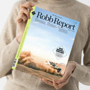 Person in beigefarbenem Strickpullover hält ein Magazin mit dem Titel „Robb Report – The New Luxury“ und der Beschriftung „Deutsche Ausgabe Nr. 38“. Das Cover zeigt ein weißes Auto, das über eine staubige Schotterstraße fährt und dabei Staubwolken aufwirbelt, vor hellem Himmel. Ein schwarzer Schriftzug im weißen Kasten lautet „Wer bremst, verliert“. Linker Rand mit hellgrünem Streifen und Barcode am unteren Ende.