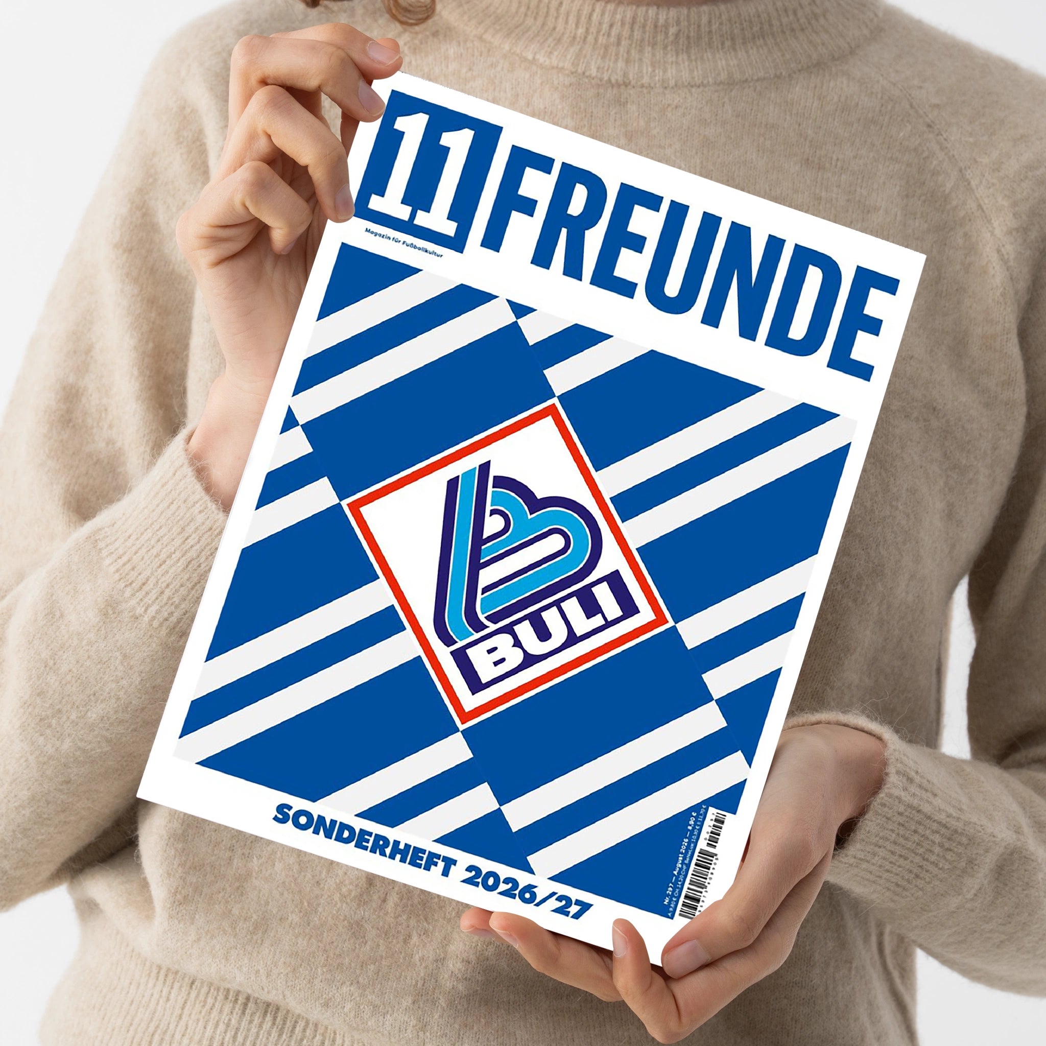 Weibliche Person im beigefarbenen Strickpullover hält ein schräg ausgerichtetes Magazin mit weißem Rand und blau-weißen Streifen. Auf dem Cover stehen „TIERFREUNDE“, „Magazin für Fußballkultur“, „BULLI“ und „SONDERHEFT 2026/27“; mittig befindet sich ein rot umrandetes BULLI-Logo.