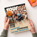 Titelbild des Magazins „TOUR“ (Ausgabe 6 | 2023) mit einem männlichen Radsportler in blauer Rennkleidung auf einem weißen Rennrad, frontal im Rennmoment aufgenommen. Textinhalte u. a.: „Europas Rennrad-Magazin Nr. 1“, „Serie Trainingsplaner – Die optimale Wettkampfvorbereitung“, „Die besten Renner“, „Radsport in der Provinz – bärenstarke Basis!“, „Hommage an den Größten“. Hände halten das Heft vor hellem Hintergrund.