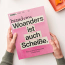 Magazin mit pinkem Umschlag, Titelaufschrift „brand eins“ und groß gesetztem Text „Woanders ist auch Scheiße. Was auf dem Arbeitsmarkt trotzdem geht“, gehalten von zwei Händen vor hellem Hintergrund; im oberen Bereich sind Barcode, Ausgabedaten und Preis klein gedruckt, rechts oben liegt ein oranger Gegenstand auf einem Teller am Bildrand.