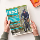 Titelblatt des Magazins „Elektro Rad“ mit blaugrünem Layout, gehalten von zwei Händen vor hellem Hintergrund. Im Mittelpunkt eine weiblich gelesene Person mit Helm und Sonnenbrille, die in einem Wald auf einem schwarzen Gravelbike fährt. Sichtbare Texte: „Flotter auf Schotter – Gravelbikes mit Spaß-Garantie“, „Der perfekte Sattel? Darauf kommt es an!“, „Rushhour-Tipps vom Verkehrs-Experten“, „EUROBIKE-NEUHEITEN: Das Beste von der Messe“.