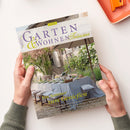 Zwei Hände halten eine Ausgabe der Zeitschrift „Garten & Wohnen Träume“ (3/2025) mit Titelbild einer mediterranen Gartenszene: gedeckter Tisch mit hellblauer Tischdecke, Stühlen, Geschirr und Gläsern unter großem Sonnenschirm, umgeben von Steinmauer, Pflanzen und weißen Blüten; sichtbare Überschriften „Ein Sommer zum Genießen“ und Themen zu mediterranen Aromen, Urlaubsideen und Gartenträumen.