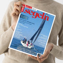 Person in hellbraunem Strickpullover hält die Zeitschrift „segeln“ (Ausgabe Mai/Juni 2025) vor neutralem Hintergrund. Das Cover zeigt ein Segelboot mit mehreren Personen auf dem Meer; weiße Yacht mit dunklem Segel und roter Flagge. Blaue Titelschrift „segeln“ oben, weitere weiße und gelbe Texte u. a.: „TOP YACHT AWARD 2025“, „NIEDERLANDE, NORDSEE, OSTSEE“ sowie Themen zu Technik, Reisen und Regatten.