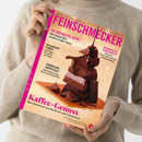 Menschliche Person in beigefarbenem Strickpullover hält das Magazin „Feinschmecker“ vor neutralem Hintergrund. Titelthemen: „Schokolade! Die Sieger des großen Tafel-Tests“, „Trendziel Tokio“, „Südtirols Aufsteiger“, „Schnitzel & Schmankerl“, „Kaffee-Genuss“. Auf dem Cover ein Stapel verschiedener Schokoladenstücke auf einem braunen Schokoteller, Farbgestaltung in Rosa, Braun und Beige.