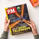 Titelbild der Zeitschrift „P.M.“ mit rotem Logo auf gelbem Hintergrund. Auf der Covergrafik ist ein großes Observatorium mit mehreren orange leuchtenden Laserstrahlen dargestellt. Textausschnitte: „NEUE SUPER-TELESKOPE“, „Exoplaneten, Gammastrahlen, Dunkle Energie“, „Bahn & KI“, „Hinter Gittern“, „Kaufe Dino“. Zwei Hände in grünen Strickärmeln halten das Magazin über einer hellen Tischoberfläche.