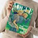 Person in beigefarbenem Strickpullover hält ein Magazin mit dem Titel „VOGUE Deutsch – Collector’s Issue“. Auf dem Cover ist eine weiblich erscheinende Person mit blondem Haar in einem geometrisch gemusterten, grün-blauen Kleid mehrfach kaleidoskopartig gespiegelt zu sehen. Text auf dem Titel: „EIN KALEIDOSKOP DES FRAUSEINS“, „THE WOMEN WE ARE“, Name unten links: „MALA EMDE“. Hintergrund in verschiedenen Grüntönen.