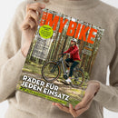 Person in beigefarbenem Strickpullover hält das Magazin „MY BIKE“ mit Titel in orangefarbenen Großbuchstaben. Auf dem Cover fährt eine Person mit Helm, roter Jacke und dunkler Hose auf einem E-Bike durch einen Wald. Textaufschriften u. a.: „Räder für jeden Einsatz“, „20 Seiten Basiswissen zum Fahrradkauf“, „Frühjahrs-Check“ und „Citytrip Lissabon“.
