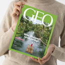 Person in beigem Wollpullover hält die Zeitschrift „GEO“ mit grünem Rand und großer weißer Titelaufschrift. Auf dem Coverfoto ist eine tropische Flusslandschaft mit dichtem Palmenbestand zu sehen, in der Mitte gleitet eine einzelne Person in roter Kleidung auf einem Bambusfloß. Weitere sichtbare Texte: „Wie Reisen uns prägt“, „Serie Hirnforschung“, „Mali“, „Diamantfieber in Afrika“, „Atomnull“.
