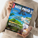 Person in beigem Strickpullover hält ein Magazin mit dem Titel „REISE & PREISE – Den Traumurlaub perfekt planen“. Auf dem Cover sind eine felsige Küste mit türkisblauem Meer, grüne Bäume und ein Strand mit Sonnenschirmen zu sehen. Sichtbare Textelemente: „Queensland – Australiens Naturwunder“, „Den Sommer verlängern“, „19 traumhafte Ziele für die Nebensaison“, sowie kleinere Reiseziele „Istrien“, „Bangkok“, „Irland“ und „Marokko“.