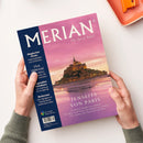 Magazin „MERIAN – The Art of Travel“ mit violett-rosa Cover, im Zentrum die Abtei Mont-Saint-Michel bei Sonnenuntergang, darunter Titel „Jenseits von Paris“ sowie Unterzeile zu Frankreichs Norden. Linke blaue Spalte mit weißen Texten zu Themen: „Magischer Oman“, „Viva Valencia!“, „Legendäre Hotelbars“ und „Bodensee“. Gehalten von zwei Händen über hellem Tisch, oben rechts eine orange Ablageschale sichtbar.