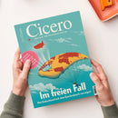Titelseite des Magazins „Cicero – Magazin für politische Kultur“ in türkis-blauer Farbgestaltung, gehalten von zwei Händen über hellem Hintergrund. Illustration zeigt eine stilisierte Weltkarte als gelb-rote Luftmatratze mit fallschirmspringender Person darunter, ergänzt durch weißen Schriftzug „Im freien Fall. Wie Deutschland sich dem Epochenbruch verweigert“ und runden pinken Aufkleber mit Text „Letztes Gefecht“.