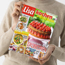 Magazin „Lisa Kochen & Backen“, Ausgabe Mai/Juni 2023, in den Händen einer Person in beigefarbenem Strickpullover gehalten. Titelbild zeigt eine Erdbeertorte mit Schokoguss auf rosa Tortenplatte, umgeben von Aufschriften wie „Himmlisch backen mit Erdbeeren“, „Extra 16 Seiten Spar-Rezepte“ und Rezeptfotos von Spargelgerichten, Suppen, Kuchen und Grillgerichten auf hellgrünem Hintergrund.
