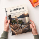 Titelseite des Magazins „Robb Report“ mit der Überschrift „Luxus der Regeneration“. Zu sehen ist eine Frau auf einem Sofa und ein Mann, der auf einer Terrasse mit Blick auf einen See und schneebedeckte Berge steht. Auf dem Glastisch davor liegen Getränke und Snacks, im Hintergrund Sonnenuntergang über dem Wasser. Oberer Rand mit Text „Design. Stil. Reise. Motoren. Werte.“ und Datumsangabe „Jul/Aug 26“.