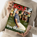 Person in beigem Strickpullover hält ein Magazin mit dem Titel „GOLF MAGAZIN“. Auf dem Cover kniet ein männlicher Golfer in weißer Hose, hellblauem Shirt und weißer Kappe auf einem Rasenplatz mit jubelnder Körperhaltung. Textaufschriften: „Das Masters“, „McIlroy holt endlich den Career Grand Slam“, „Südtirol“, „Trouble-Shots“, „Im Test“, „Les Bordes“, „Ryder Cup“. Hintergrund zeigt unscharfe Menschenmenge auf Golfplatz.