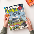Titelblatt des Magazins „Camping & Reise“, Ausgabe 03–25 Frühling. Im Mittelpunkt steht ein weißes Wohnmobil auf einer Wiese mit einer stehenden weiblichen Person daneben unter blauem Himmel. Überschriften u. a.: „Die besten Frühlingstouren in Deutschland und weltweit“, „Glamping in Kärnten“, „Magisches Trentino“, „Süddeutschland“, „Teilintegrierte – die neuesten Fahrzeuge“. Hände halten das Magazin über hellem Tisch mit oranger Schale.