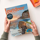 Titelbild der Zeitschrift „outdoor – Wandern. Reisen. Abenteuer.“ mit großem orangefarbenem Logo, gehalten von zwei Händen auf hellem Hintergrund. Abgebildet ist eine felsige Küste bei Sonnenuntergang mit einer stehts auf dem Klippenrand stehenden Person. Textelemente: „Die schönsten Küstentouren“, „Peaks of the Balkans“, „Testsieger: Die besten Produkte 2026“, „Zelte im Sturm“. Unten rechts ein blaues Zeltfoto.