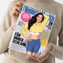 Titelblatt der Zeitschrift „Women’s Health“ mit einer lächelnden dunkelhaarigen weiblichen Person in gelbem Cropped-Hoodie und blauen Radlerhosen auf hellgrauem Hintergrund. Sichtbare Aufschriften: „FÜR IMMER SCHLANK“, „28 Tricks, besser zu essen“, „Effektiv trainieren ohne Gym“, „Dein Weg zum Super-Bauch“. Logo „FABLETICS“ auf dem weißen Bund des Tops, obere Zeile „STRONGER EVERY DAY“.