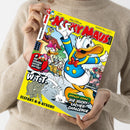 Person in beigefarbenem Strickpullover hält ein Comicheft mit dem Titel „Walt Disney Micky Maus Magazin“. Auf dem farbenfrohen Cover ist eine große Zeichnung von Donald Duck in blau-gelber Matrosenkleidung zu sehen, daneben weitere Figuren in einer Gartenszene. Mehrere Aufschriften: „+ Die große Witz-Battle!“, „Brüller-Comic: Die Nicht-Lachen-Challenge“, „W.I.T.T. – Elefant-H-H-Atschi!“, Hintergrund in Gelb und Hellblau gestaltet.