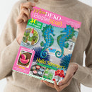 Person in beigem Strickpullover hält eine Ausgabe der Zeitschrift „Mein DEKO- und Bastelspaß“ mit pinkem Titelbalken. Auf dem Cover zwei blaue Seepferdchen vor türkisblauem Hintergrund, Aufschrift „Hallo Sommer!“ sowie Hinweise auf „30 tolle DIY-Projekte“, „Selbst gemacht: Fruchtige Sommerdekoration“ und Themen wie Gartenzwerg, Bastelfrosch und maritime Bastelideen, in bunten Farbtönen gestaltet.