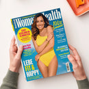 Titelbild des Magazins „Women’s Health“ mit einer lachenden Frau in einem gelben Badeanzug vor hellblauem Hintergrund. Textinhalte u. a.: „Easy schlank in 15 Minuten“, „Lebe fit & happy“, „100 % Sommer“, „Glow-Guide für schöne Haut“, „Gesundes BBQ leicht gemacht“, „Die schönste Beachwear für jede Figur“ und „Love-Check – Passt ihr gut zusammen“. Hände halten das Magazin über einer hellen Tischfläche mit orangefarbenem Objekt am Rand.