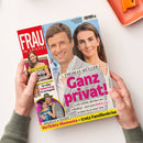 Titelblatt der Zeitschrift „Frau im Spiegel“, gehalten von zwei Händen über hellem Tisch. Im Mittelpunkt ein männlicher und eine weibliche Person vor blauem Hintergrund, daneben pinke Überschrift „Ganz privat!“ und Name „Thomas Müller“. Weitere Textelemente: „Ehefrau Lisa“, „Verliebte Momente – trotz Familienkrise“, „Ana Ivanovic – Allein in Paris!“, „Meghan & Harry“, „Ihr stiller Geburtstag in Los Angeles“. Layout mit roten, gelben und weißen Bereichen.