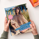 Titelblatt der Zeitschrift „Grazia“ mit einer weiblich gelesenen Person in hellbeiger, locker fallender Bluse und Sandalen, sitzend auf einem sandigen Boden vor hohem Gras. Sichtbare Textelemente: „HIGH JEWELLERY ISSUE“, „ANZIEHEN & SENSATIONELL AUSSEHEN“, „DIE 50 SCHÖNSTEN SOMMERKLEIDER“, „DER STYLE-GUIDE ZUR FUSSBALL-WM: JEDER LOOK EIN TREFFER“, „MOST WANTED“. Hände halten das Magazin über einer hellen Oberfläche mit orangener Ablage.