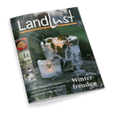 Titelbild der Zeitschrift „Landlust“, Winterausgabe mit orangefarbenem Balken unter dem Logo und dem Heftthema „Winterfreuden“. Abgebildet sind zwei Eislaternen mit eingeschlossenen grünen Blättern und roten Beeren auf einem dunklen Holztisch im Freien. Links am Rand drei Fotoelemente mit den Themen „Herzhafte Eintöpfe“, „Wildgänse“ und „Selbstgemachte Figuren“. Weitere sichtbare Texte: „Beim Schneeglöckchen-Sammler“, „Lichter & Laternen für ein warmes Leuchten“, „Auf die Schlitten: Schöne Naturpisten“.