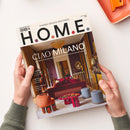 Zwei geschlechtsneutral erscheinende Hände halten das Magazin „H.O.M.E.“ mit dem Titeltext „CIAO MILANO – Das Beste vom Salone del Mobile und der Milan Design Week“. Auf dem Cover ist ein elegantes Wohnzimmer mit violett-goldener Wandgestaltung, roten Sofas, runden Hockern in Orange und Grau, einem roten Tisch und einem goldenen Kronleuchter zu sehen, vor einem hellen Hintergrund mit Türöffnung und Spiegel.