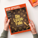 Titelseite des Magazins „DER SPIEGEL“ mit orangefarbenem Rahmen, der Titel lautet „Die Luxus-Falle – Wie Hermès, Rolex und Co. uns manipulieren“, abgebildet auf einer braunen Lederstruktur mit goldenen Luxusmarken-Symbolen wie LV, Krone, Totenkopf und Schlange; gehalten von zwei hellhäutigen Händen über einer weißen Oberfläche mit orangefarbenem Tablett im Hintergrund.