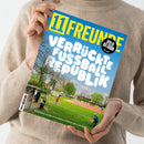 Person in beigefarbenem Strickpullover hält das Magazin „11 FREUNDE“ mit gelbem Logo auf blauem Hintergrund. Titeltext in weißen Großbuchstaben: „Verrückte Fußball Republik“. Darunter ein Foto eines kleinen Fußballplatzes mit Spieler*innen in roten Trikots, Zuschauer*innen am Spielfeldrand und grünen Bäumen im Hintergrund. Schwarzer Kreis oben rechts mit weißer Aufschrift „Spieler der Saison“, unten Textzeile: „Eine Reise zu den kuriosesten Plätzen des Landes“.
