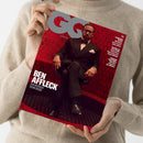 Eine hell gekleidete Person hält ein „GQ Germany“-Magazin mit dunkelrotem Cover und sechseckigem Fliesenmuster; auf dem Titel sitzt ein männlich wirkender Mann im schwarzen Anzug, weißem Hemd und Fliege mit überkreuzten Beinen auf einer Bank. Sichtbare Textelemente: „BEN AFFLECK – EIN BESUCH BEIM BOSS“ sowie kleinere Schlagzeilen zu weiteren Namen und Themen am rechten Rand.