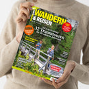 Titelblatt des Magazins „Wandern & Reisen 02·25 Frühling“, gehalten von einer Person in beigem Pullover vor neutralem Hintergrund. Das Cover zeigt einen Mann und eine Frau mit Rucksäcken auf einer Holzbrücke in grüner Berglandschaft. Sichtbare Texte: „32 Traumtouren für die schönste Jahreszeit“, „Ursprüngliches Graubünden“, „Ausrüstungsratgeber“, „Steiermark genießen“ sowie ein oranger Kreis mit der Aufschrift „EXTRA Wanderreisen mit Veranstalter“.