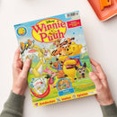 Kinderzeitschrift „Disney Winnie Puuh“ mit bunter Illustration der Comicfiguren Winnie Puuh, Tigger, Ferkel und Rabbit auf einer Wiese, umgeben von Bäumen und Vögeln. Sichtbare Texte: „EXTRA: Shopping-Set 8 Teile“, „Poster und Rätsel“, „Naturforscher – süßer Tiernachwuchs“, „Zum Herausnehmen!“, „Entdecken  Malen  Spielen“. Gehalten von zwei Händen vor hellem Hintergrund mit orangefarbenem Tablett.