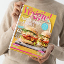 Person in beigefarbenem Strickpullover hält eine Zeitschrift mit dem Titel „slowly veggie! Vegetarisch & vegan genießen“. Auf dem gelb‑rosa Cover sind zwei üppig belegte Burger mit Sesambrötchen, Kraut und Gemüse vor hellem Hintergrund zu sehen. Text auf dem Titel: „Burger, Wraps & Bowls“, „68 Gute‑Laune‑Rezepte für den Sommer“, „Beren‑Saison“, „Easy Streetfood für jeden Tag“, „Coolcation“ und „Smokey Jackfruit‑Burger mit Coleslaw“.