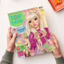 Titelblatt des Magazins „TOPModel by Depesche“ mit einer gezeichneten weiblichen Figur mit langen, hellblonden Haaren und pinkem Pullover, die mit den Händen ein Herz formt. Der Hintergrund zeigt Farbverläufe in Rosa und Gelb, umliegende Textfelder mit Überschriften wie „Knusper-Frühstück“, „Ein unvergesslicher Geburtstag“, „Happy Ostern“ und „Stylecheck“. Zwei Hände halten das Heft über einer weißen Oberfläche.