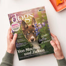 Titelseite der Zeitschrift „dogs – Mit Hund leben“, Ausgabe 3/2025 Mai | Juni. Abgebildet ist ein braun-weißes Hundejunges mit blauen Augen vor violett-grünen Blumen. Sichtbare Schlagzeilen: „Von Machos und Zicken – Vierbeinige Persönlichkeiten im Rausch der Hormone“, „Perfekter Garten für Vierbeiner“, „Spezial Tierarzt – Kosten, Stress & Notfälle“, „Fürs Grüne“, „Pro & Contra“, „Checkliste Europa“. Gehalten von zwei Händen über hellem Untergrund.