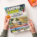 Titelbild der Zeitschrift „Camping & Reise“ mit der Überschrift „Der perfekte Sommerurlaub in Deutschland und Europa“. Im Mittelpunkt ein Foto eines Wohnmobils auf einer kurvigen Landstraße durch hügelige Landschaft, dazu Textfelder mit Themen wie „Luxus-Reisemobile“, „Portugal“, „Kärnten entdecken“, „Schweiz“, „Bretagne“ und „Vorzelt“. Gehalten von zwei Händen über heller Tischoberfläche mit orangefarbenem Tablett.