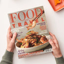 Hände einer Person in grünem Strickpullover halten das Magazin „Food and Travel Deutsch“. Das Titelbild zeigt eine Schale mit gebratenen Garnelen, eine Garnele wird mit Essstäbchen darüber gehalten. Aufschriften u. a.: „ASIATISCHE KÜCHE“, „REZEPTE VON DER DONAU“, „SPEKTAKULÄRE SPAS“, „City Guide MÜNCHEN“ und „FOODIE-HOTSPOTS – Städte zwischen Streetfood und Sternen“.