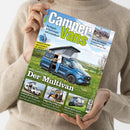 Person in beigem Wollpullover hält Magazin „CamperVans – Das Magazin für Kastenwagen und Campingbusse“ in beiden Händen, Titelbild zeigt blauen Campervan mit geöffneter Seitentür und aufgestelltem Hochdach auf einer Wiese, ein männlicher Mensch steht an der Tür, Aufschriften u. a. „Der Multivan“, „Fahrwerk-Upgrade“, „Tour de France“, „Forster für Fünf“, „Sprinter ohne 4x4“ und „Mehr geht nicht“.