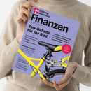 Person in beigem Strickpullover hält eine Zeitschrift mit hellviolettem Titelblatt. Oben links befindet sich das rot-weiße Logo „Stiftung Warentest“, daneben der Titel „Finanzen“. Hauptüberschrift: „Top-Schutz für Ihr Rad“. Abgebildet ist ein gelbes Fahrrad mit schwarzem Sattel, silbernem Faltschloss und schwarzem Reifen. Weitere Texte thematisieren u. a. Börsenturbulenzen, Steuern, Immobilien und Versicherungen.