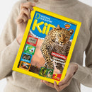 Helle Zeitschrift „National Geographic KiDS“ mit gelbem Rand, mittig ein Leopard auf beigem Hintergrund, darüber blauer Schriftzug mit Logo. Seitlich und unten mehrere kleinere Bilder von Tieren mit Texten wie „GEPARD: keiner rennt schneller!“ und „DSCHUNGEL: Leoparden im Regenwald!“. Eine hell gekleidete, langärmlige Person hält das Magazin vor neutralem Hintergrund in beiden Händen.