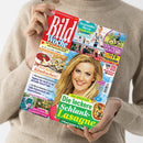 Person mit beigem Strickpullover hält das farbenfrohe Cover der Zeitschrift „Bild Woche“. Titelthemen: „Wir lieben Käsekuchen“, „Die leckere Schlank-Lasagne – 4 Pfund leichter in 3 Tagen“, „Tap­pen Sie nicht in die aktuelle Anruf-Falle“ und Artikel zu Gesundheit, TV-Programm und Freizeit. Zentral ein Porträt einer lächelnden blonden Frau vor hellblauem Hintergrund, flankiert von kleineren Bildern zu Essen, Natur und Fitness.