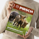 Person in beigem Strickpullover hält die Jagdzeitschrift „Pirsch“ mit braunem Titelbalken und weißer Aufschrift „PIRSCH – Respekt vor dem Wilden. Seit 1879.“; Ausgabe 11/2025. Auf dem Cover steht vorne ein junges Wildschwein auf grünem Gras, unscharf im Hintergrund weitere Frischlinge. Textausschnitte: „Rehböcke“, „Aufbrechsägen“ und „Sommersauen – Das müssen Sie bei der Nachtjagd beachten“.