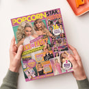Magazin „Popcorn Star & Poster“ mit pink-gelbem Titelbalken, zentral zwei weibliche Personen mit langen Haaren in glamouröser Kleidung, verschiedene kleinere Porträtbilder von Stars in farbigen Kästchen, gelbe und pinke Schlagzeilen wie „Fake or Fact?“ und „16 coole Poster“, Textbereich unten mit Themen zu Kino, Quiz, News und Starhoroskop, gehalten von zwei Händen über hellem Untergrund.
